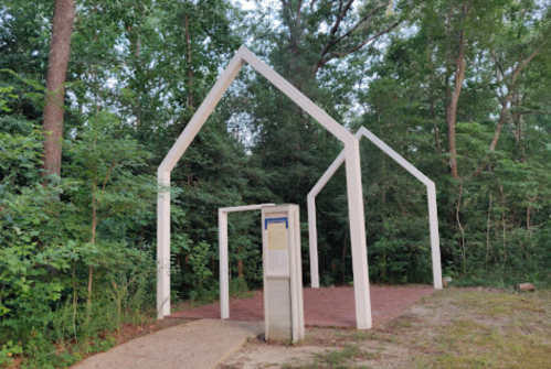 A minimalist white structure resembling a house frame, surrounded by trees and greenery, with an informational sign nearby.