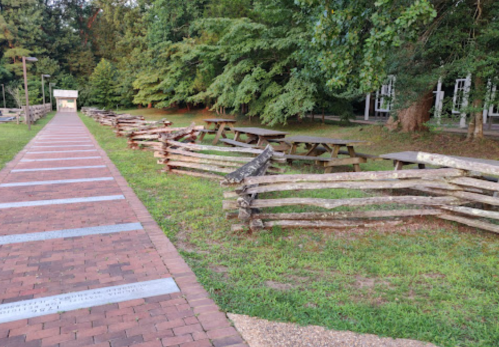 A brick pathway leads to picnic tables surrounded by rustic wooden fences and greenery in a park setting.