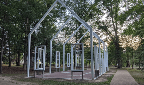 A skeletal white structure resembling a house, surrounded by trees, with open windows and a pathway nearby.
