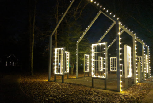 A lit structure resembling a house frame, adorned with string lights and surrounded by trees at night.