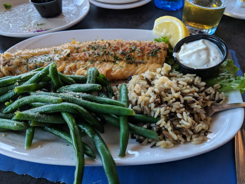 A plate with grilled fish, green beans, wild rice, a lemon wedge, and a small cup of sauce.