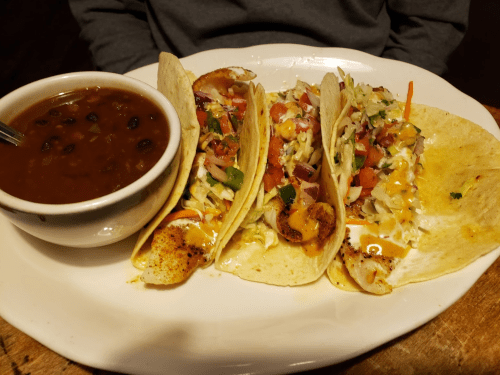 Three tacos filled with colorful toppings, served with a side of black beans in a bowl.