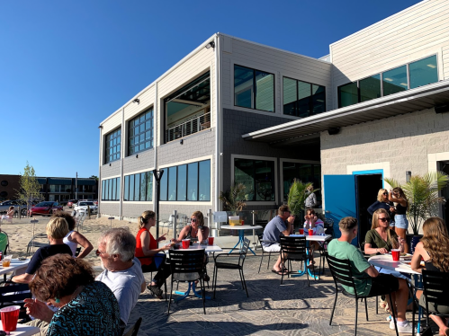 A lively outdoor dining area with people enjoying meals at tables, set against a modern building and clear blue sky.