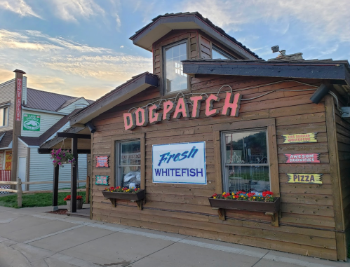 A wooden building with "Dogpatch" sign, featuring a fresh whitefish banner and colorful flower boxes.