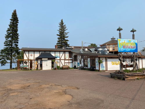 A rustic restaurant with a sign, surrounded by trees and a gravel parking area, near a body of water.