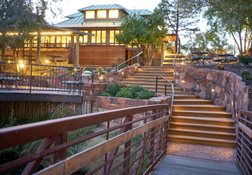 A wooden bridge leads to a cozy, illuminated building surrounded by trees and landscaped steps at dusk.