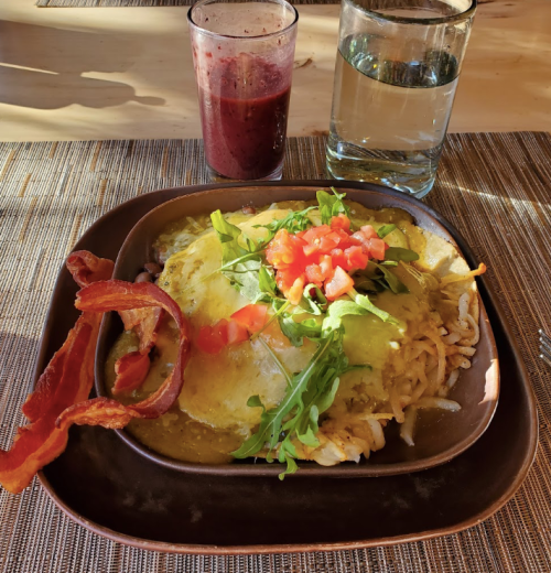 A plate of breakfast with eggs, bacon, hash browns, topped with tomatoes and greens, alongside a smoothie and water.