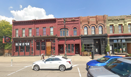 Historic brick buildings with storefronts, including a "NOW OPEN" sign, and parked cars in front on a sunny day.