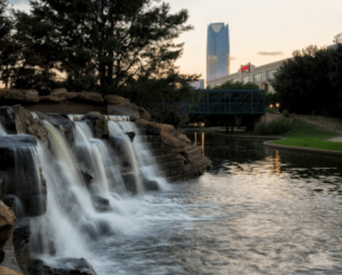 A serene waterfall flows into a calm pond, with a city skyline in the background at sunset.