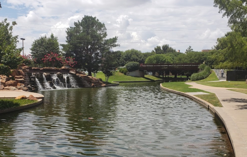 A serene park scene featuring a winding pond, a waterfall, and a wooden bridge surrounded by lush greenery.