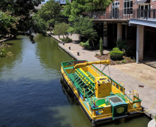 A yellow boat with a crane is docked along a calm, green canal lined with trees and buildings.