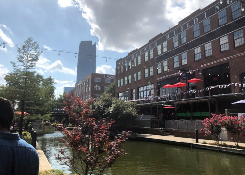 A scenic view of a canal lined with trees and buildings, featuring outdoor seating and a blue sky with clouds.