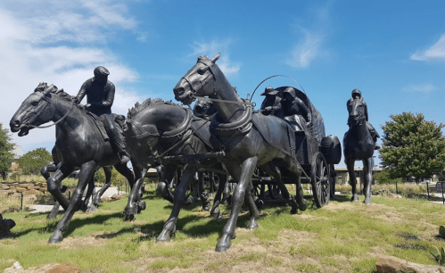 A bronze sculpture of horses and riders pulling a wagon, set against a blue sky and green grass.