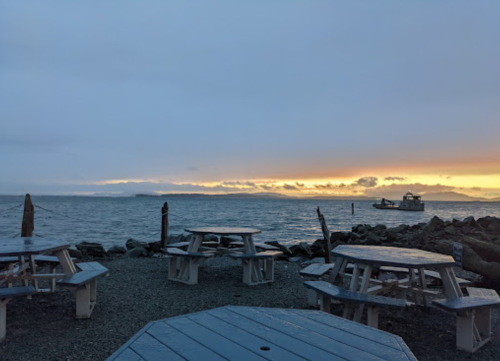 A serene waterfront scene at dusk, featuring picnic tables and a boat on the water against a colorful sky.