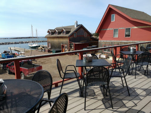 View of a waterfront restaurant patio with black tables and chairs, overlooking a harbor with boats and red buildings.