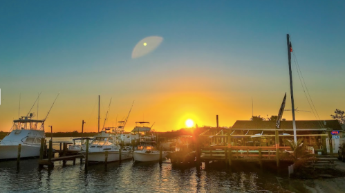 Sunset over a marina with boats docked and a restaurant, casting warm orange and yellow hues across the sky.