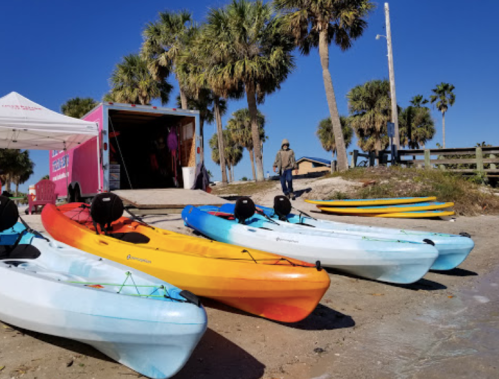 Colorful kayaks lined up on a sandy beach with palm trees and a trailer in the background under a clear blue sky.
