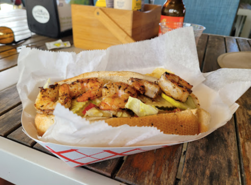 A shrimp sandwich with grilled shrimp, lettuce, pickles, and sauce, served in a paper-lined basket on a wooden table.