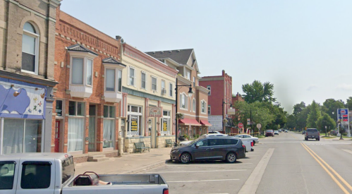 A small town street lined with brick buildings, shops, and parked cars under a clear blue sky.