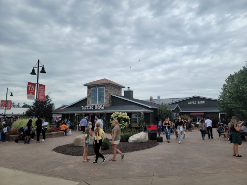 A bustling outdoor area with a tasting room and apple barn, surrounded by people and cloudy skies.