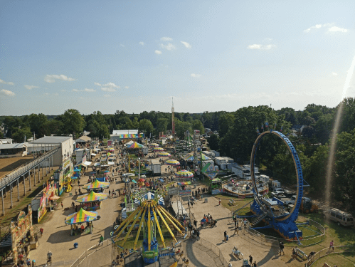 Aerial view of a vibrant fairground with rides, games, and colorful tents under a clear blue sky.