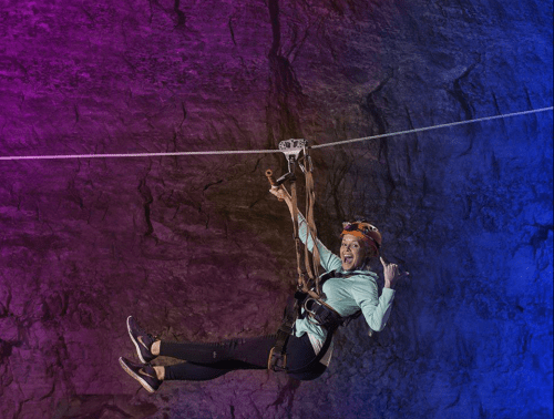 A person zip-lining through a cave, wearing a helmet and harness, with a joyful expression against a colorful background.