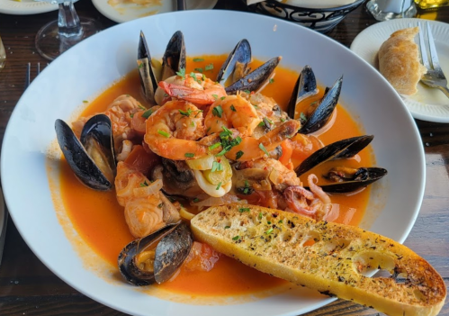 A bowl of seafood pasta with shrimp, mussels, and a slice of garlic bread, garnished with parsley in a rich tomato sauce.