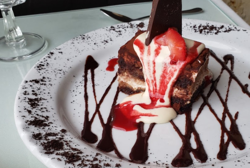 A slice of chocolate dessert topped with strawberries and drizzled with cream and chocolate sauce on a white plate.