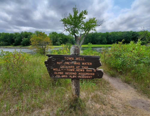 A wooden sign near a pond reads "Town Well" with historical notes about water and a resident named Mrs. Alby.