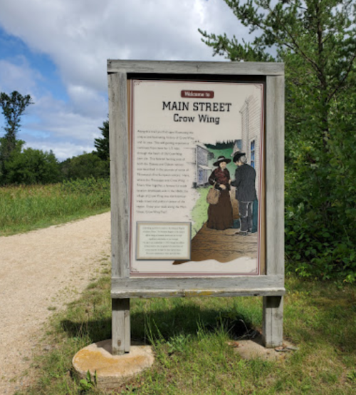 Sign welcoming visitors to Main Street, Crow Wing, with historical illustrations and text about the area. Greenery in background.