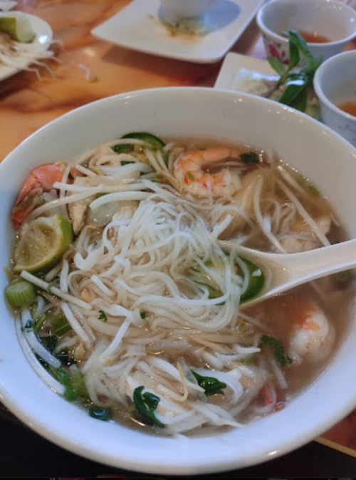 A bowl of noodle soup with shrimp, herbs, and lime slices, served with a spoon.