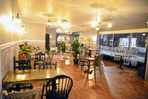 Cozy café interior with wooden tables, chairs, and plants, featuring large windows and warm lighting.
