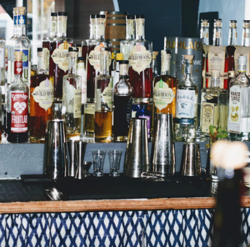 A bar shelf filled with various bottles of liquor and cocktail tools, with a patterned cloth in the foreground.