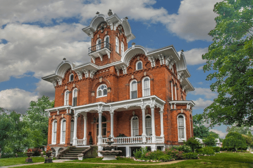 A grand, red brick Victorian mansion with ornate details, large windows, and a tower, set against a cloudy sky.