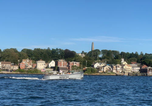 A water taxi moves through the water near a shoreline with houses and a tall monument in the background under a clear blue sky.