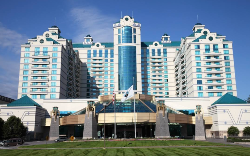 A large, modern hotel with a glass facade and turquoise accents, surrounded by green lawns and blue skies.