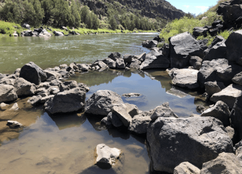 A serene river scene with smooth rocks and calm water reflecting the surrounding greenery and rocky landscape.