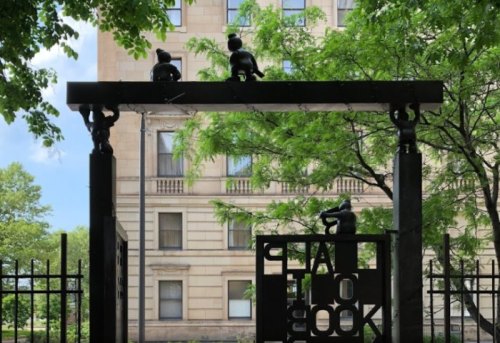 A metal archway with sculptures of children, surrounded by trees and a building in the background.