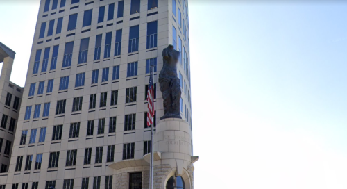 A tall building with a statue of a figure on a pedestal, alongside an American flag. Clear blue sky in the background.