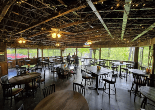 A rustic restaurant interior with wooden tables, chairs, and string lights, featuring a view of greenery outside.