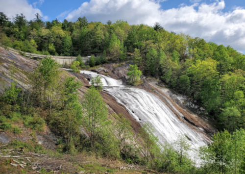 A cascading waterfall flows down a rocky slope, surrounded by lush green trees under a partly cloudy sky.