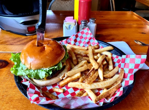 A burger with lettuce and cheese, topped with a knife, served with a side of golden fries in a red-checkered basket.