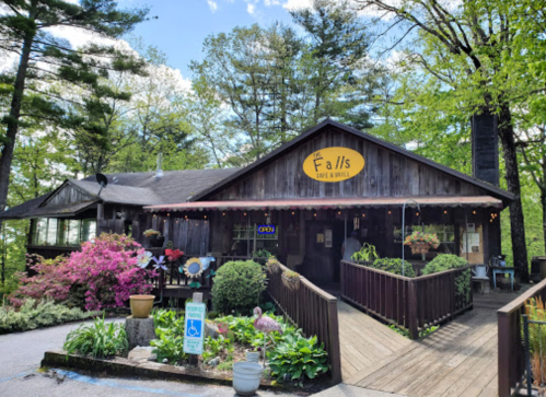 A rustic restaurant with a wooden exterior, surrounded by greenery and flowers, featuring an "Open" sign.