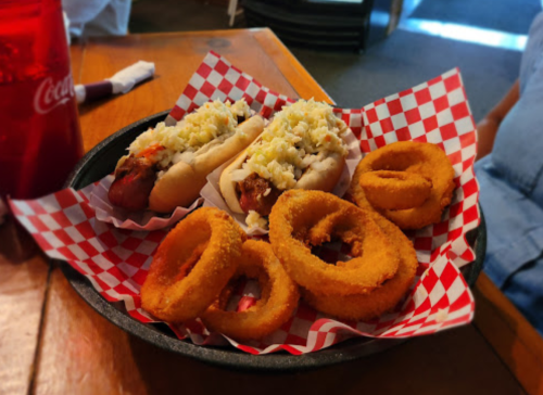 A plate with two hot dogs topped with coleslaw and a side of crispy onion rings, served in a red-checkered basket.