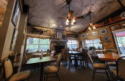 Cozy restaurant interior with wooden tables, chairs, and a stone fireplace, adorned with rustic decor and large windows.