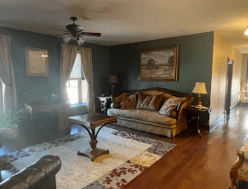 Cozy living room with a sofa, decorative pillows, a coffee table, and warm lighting, featuring dark green walls and artwork.