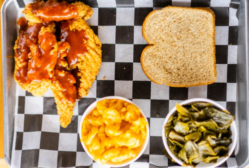 A tray with fried chicken tenders in sauce, a slice of bread, macaroni and cheese, and collard greens on a checkered background.