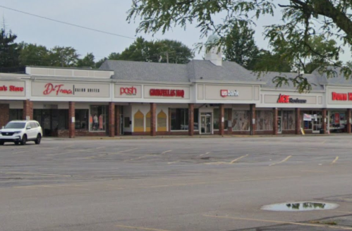 A row of storefronts in a shopping plaza, featuring various businesses and a parking lot in front.