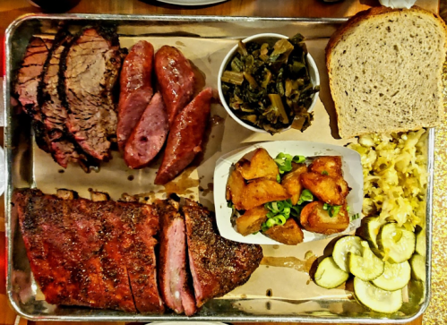 A metal tray filled with various meats, sides of greens, potatoes, pickles, and slices of bread.