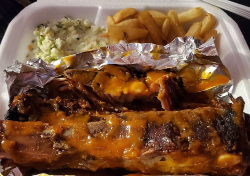A takeout container with BBQ ribs covered in sauce, coleslaw, and French fries on the side.